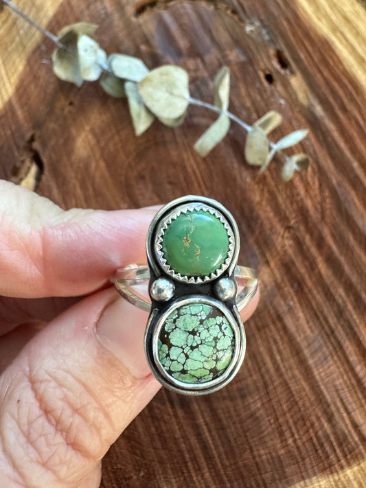 Hand holding a silver ring with green stones on a wooden surface